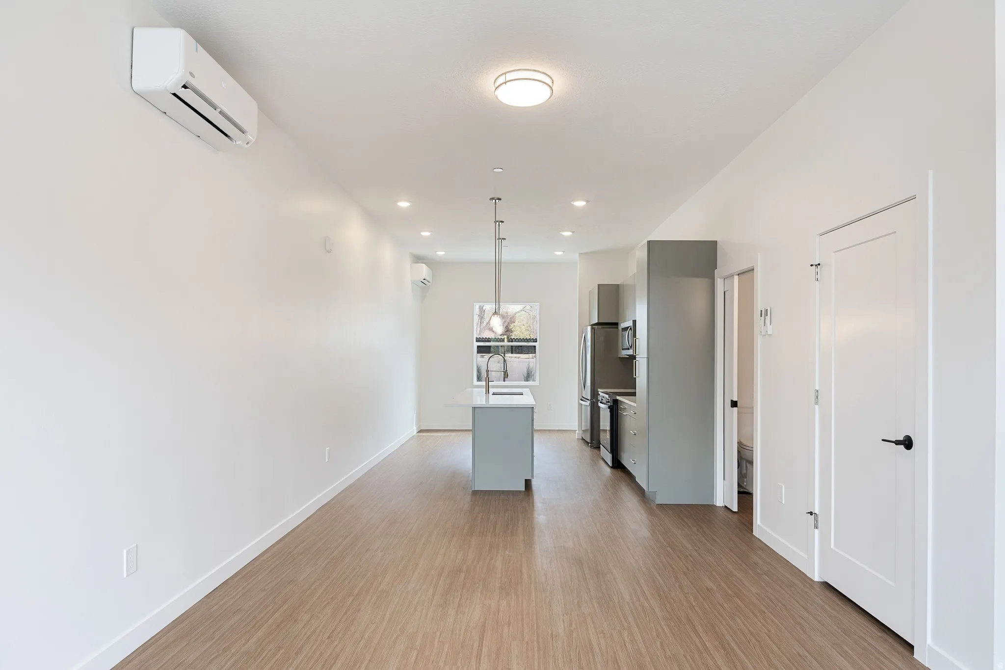 Open-concept living area with wood-finish flooring, a kitchen island with pendant lighting, stainless steel appliances, and wall-mounted climate control units