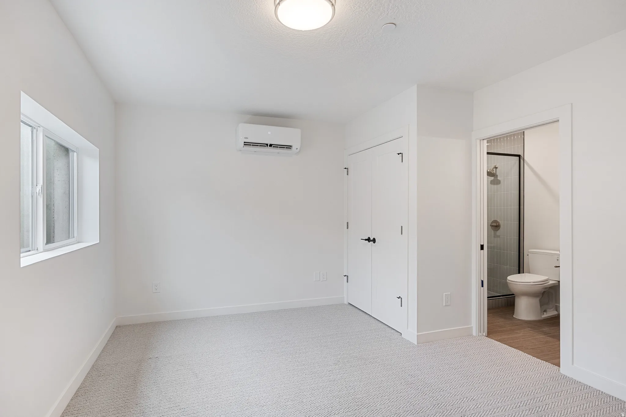 Recessed ceiling lighting, wall-mounted air conditioning unit, white interior doors, window with white trim, and light-colored patterned carpet