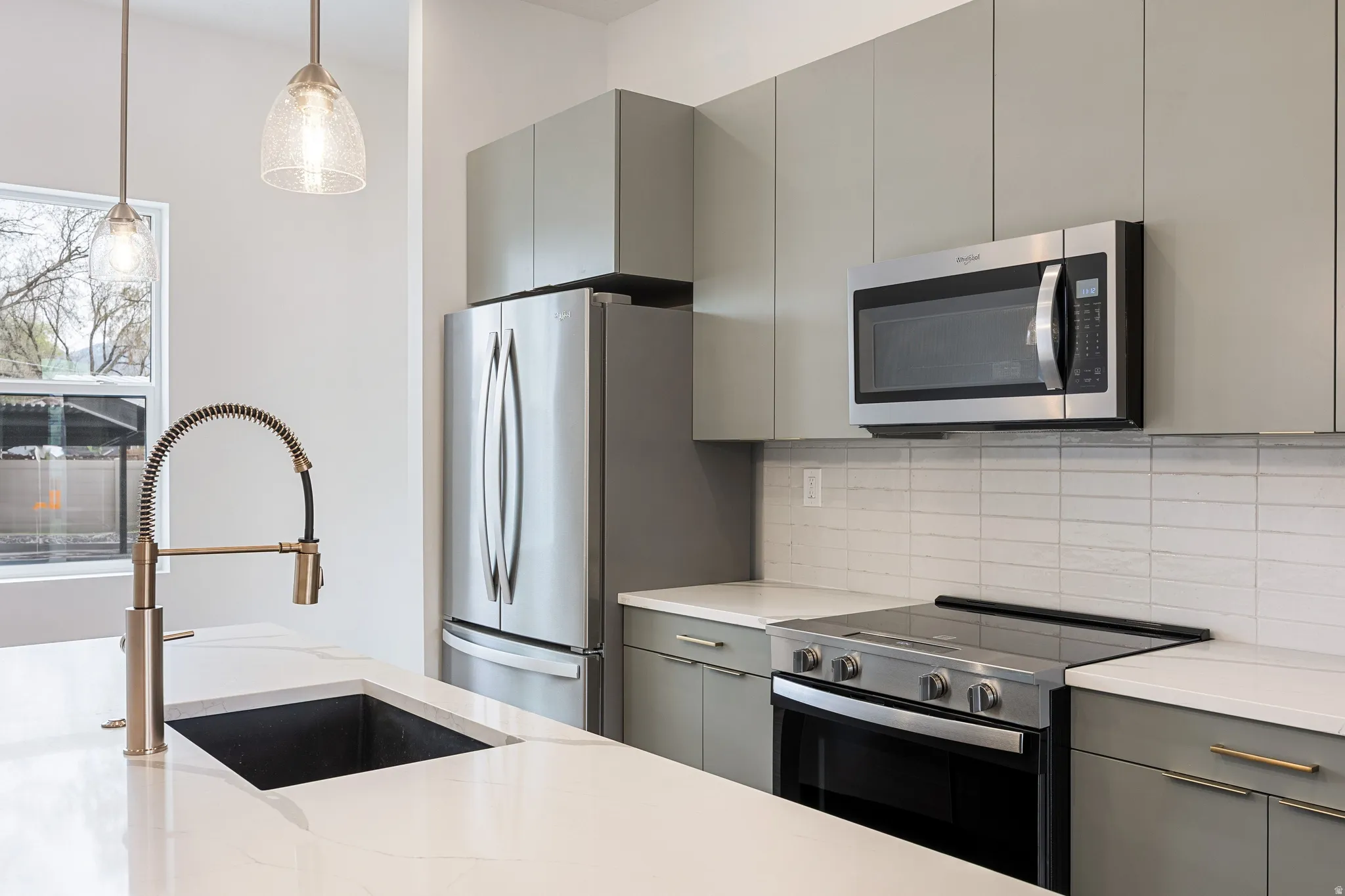 Contemporary kitchen featuring gray flat-panel cabinetry, stainless steel appliances, white tile backsplash, and a large kitchen island with an integrated sink and brushed nickel faucet