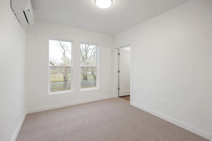 Crisp white walls, two single-hung windows, light-colored patterned carpeting, a white overhead flush-mount light fixture, and a wall-mounted mini-split unit