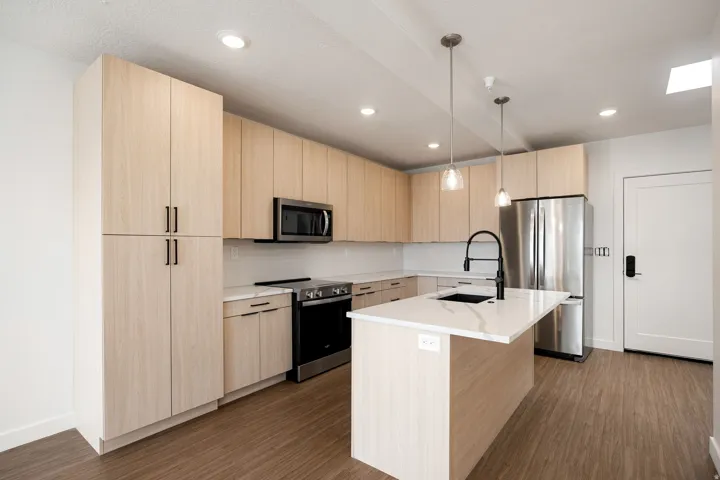Kitchen featuring light wood-finish cabinetry, stainless steel appliances, a center island with a sink, pendant lighting, and wood-finish flooring