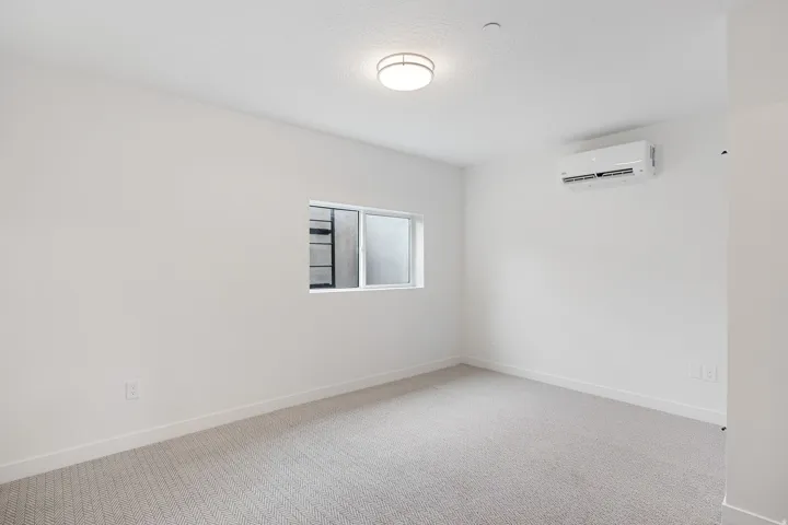 White walls, gray patterned carpeting, flush mount ceiling light, horizontal sliding window, and a wall-mounted climate control unit
