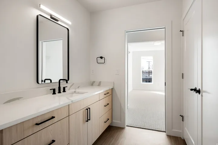 White countertop with integrated sink, black fixtures, wood-finish cabinetry, and contemporary mirror with overhead lighting