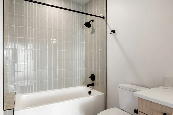 Bathtub and shower combination with vertical stacked tile surround, matte black shower fixtures, and a light wood-finish vanity with a light countertop