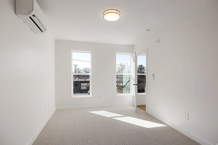White walls, two windows, wall-mounted air conditioning unit, ceiling-mounted light fixture, and light-colored carpet