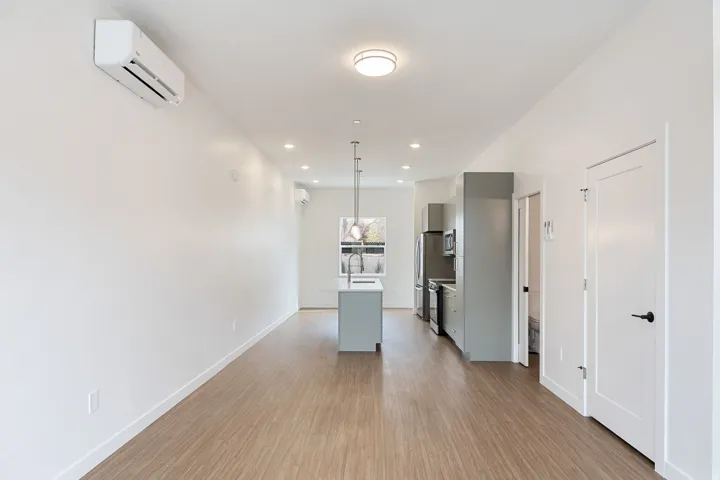 Open-concept living area with wood-finish flooring, a kitchen island with pendant lighting, stainless steel appliances, and wall-mounted climate control units