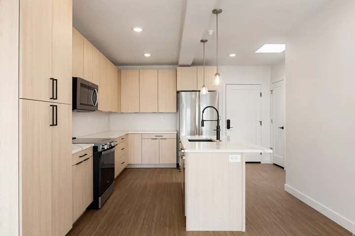 Modern kitchen with light wood-finish cabinetry, stainless steel appliances, white countertops, and wood-finish flooring