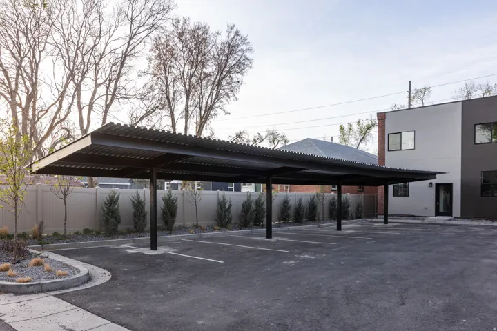 Asphalt parking area with multiple covered parking spaces, a contemporary building exterior with dark and light gray siding, and a privacy fence