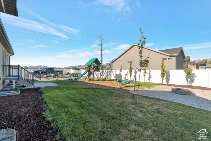 Grassed yard area featuring a gravel patio, stone fire pit, wood chip landscaping, and a white privacy fence