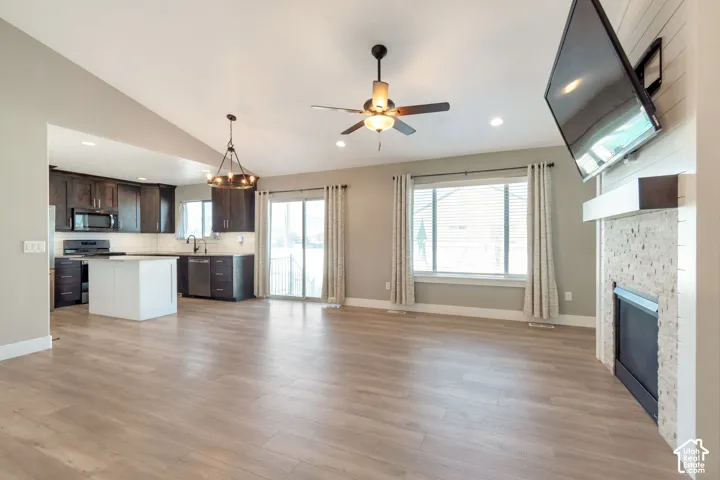 Vaulted ceiling with a ceiling fan, wood-finish flooring, a kitchen with dark cabinetry and a white island, a fireplace with a textured surround, and a sliding glass door