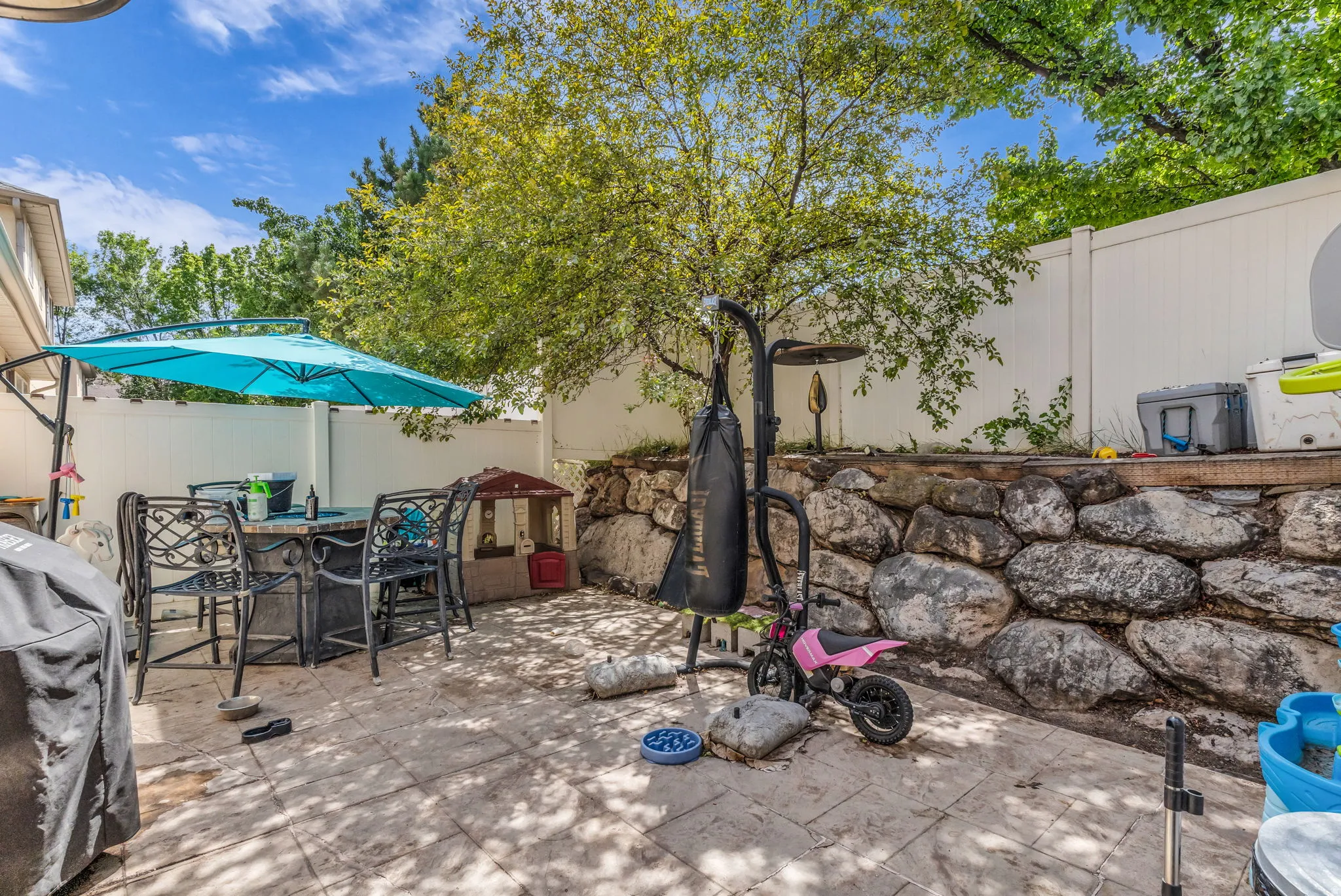 Paved patio area featuring a stone retaining wall, mature trees, and a privacy fence