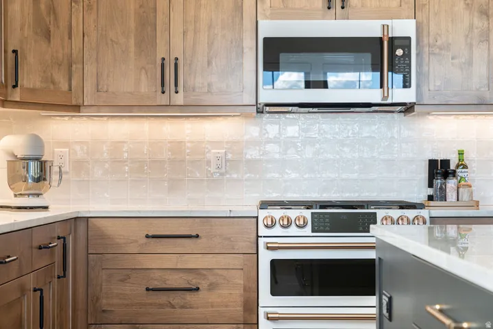 Light wood-finish cabinetry with black hardware, white tile backsplash, under-cabinet lighting, and stainless steel microwave