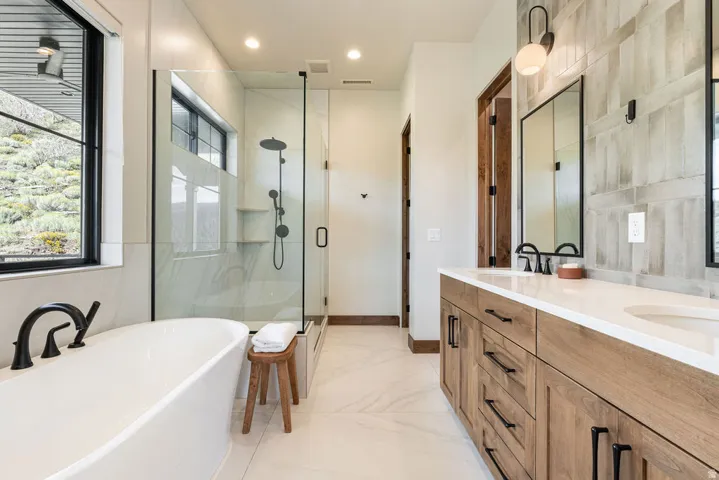 Freestanding soaking tub with matte black fixtures, glass-enclosed shower, dual vanity with wood-finish cabinetry, and white countertops