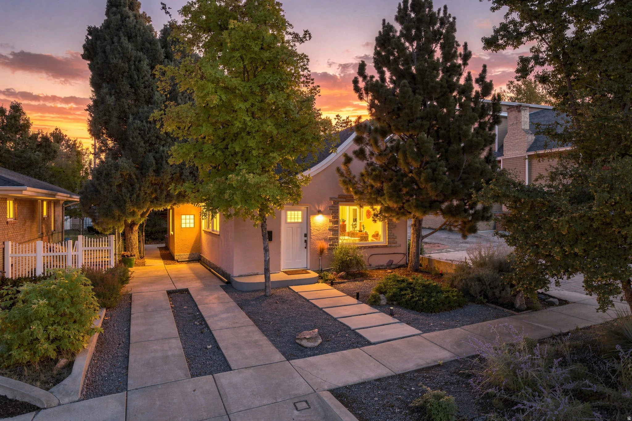 Stucco exterior with stone accents, a concrete walkway, established landscaping, and a white picket fence