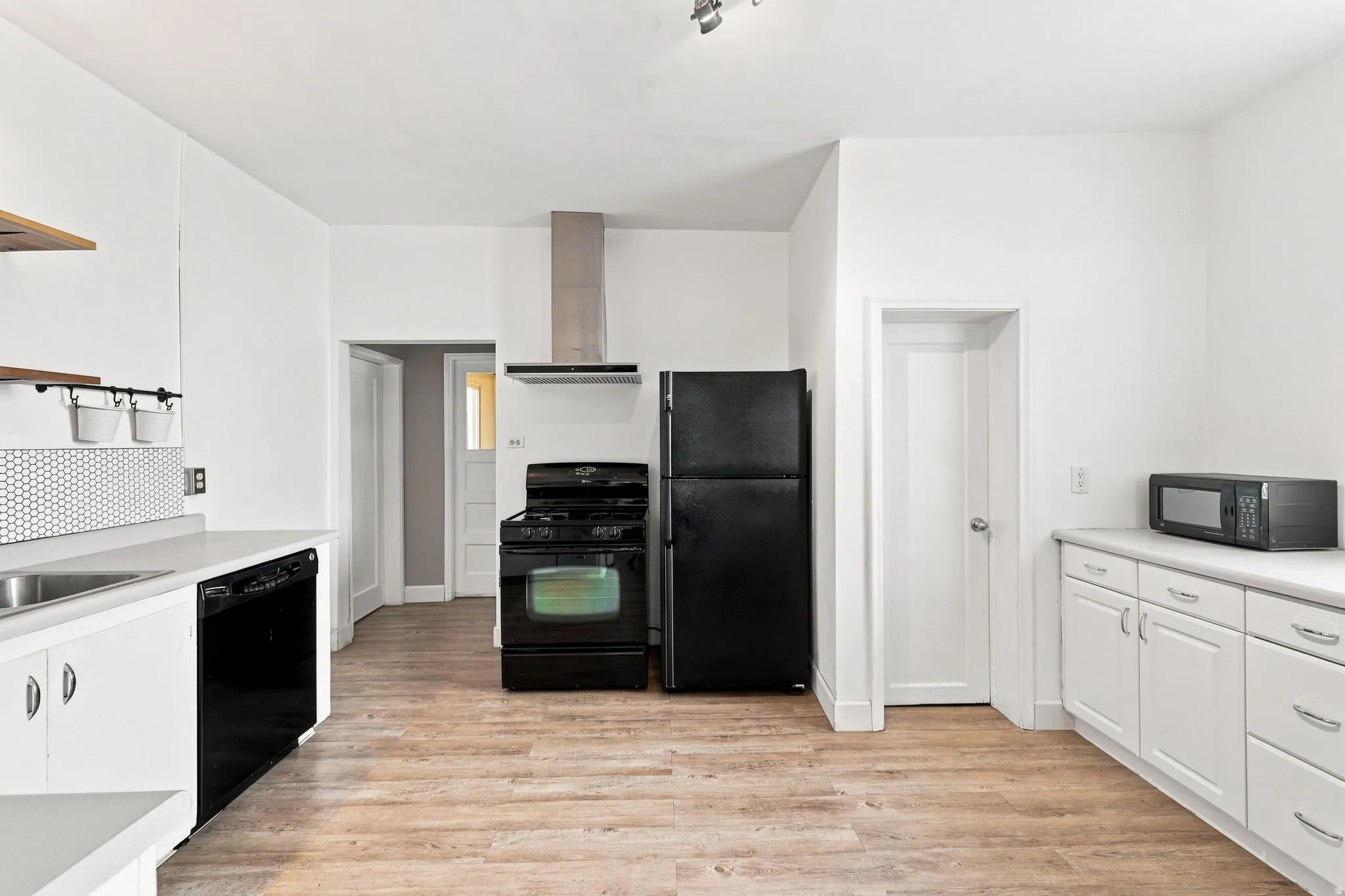 Kitchen featuring wood-finish flooring, white cabinetry, a hexagonal tile backsplash, stainless steel range hood, and a black appliance suite