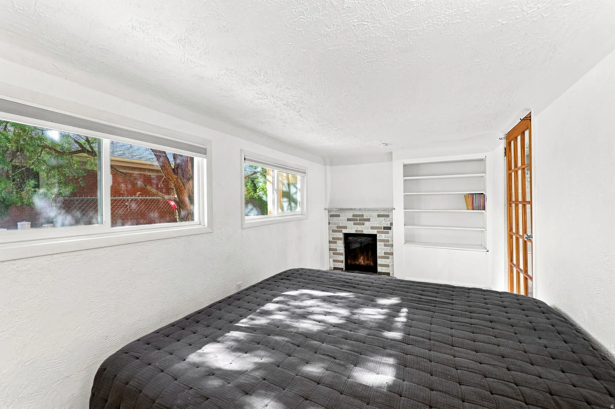 Bright, light-filled room showcasing black tile flooring with geometric pattern, large windows framing natural outdoor scenery, white walls and ceiling, a fireplace with tile surround and built-in shelving, and a wood-frame glass door providing access to