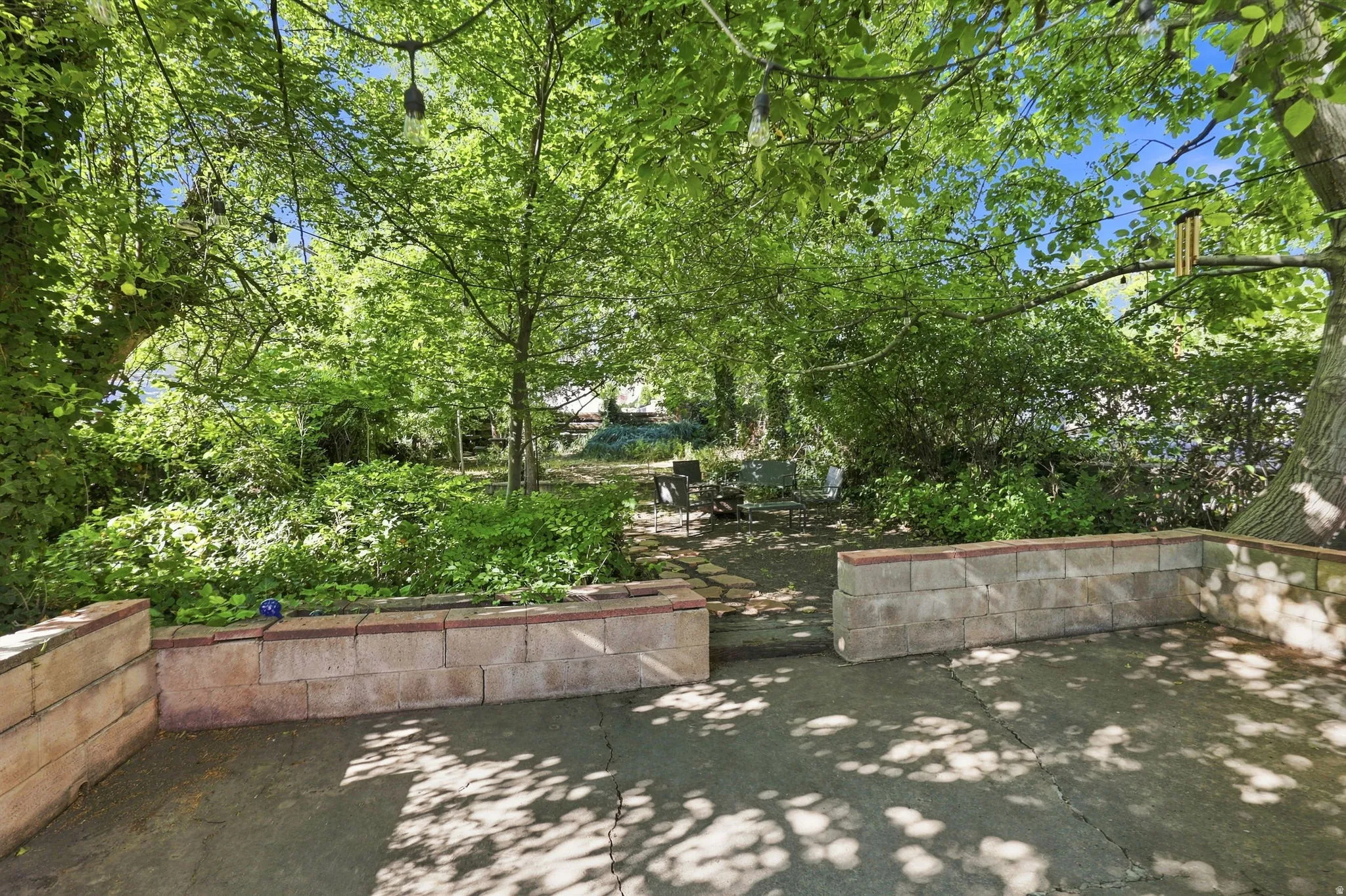 Paved patio area, raised garden beds with block construction, mature trees, and string lighting
