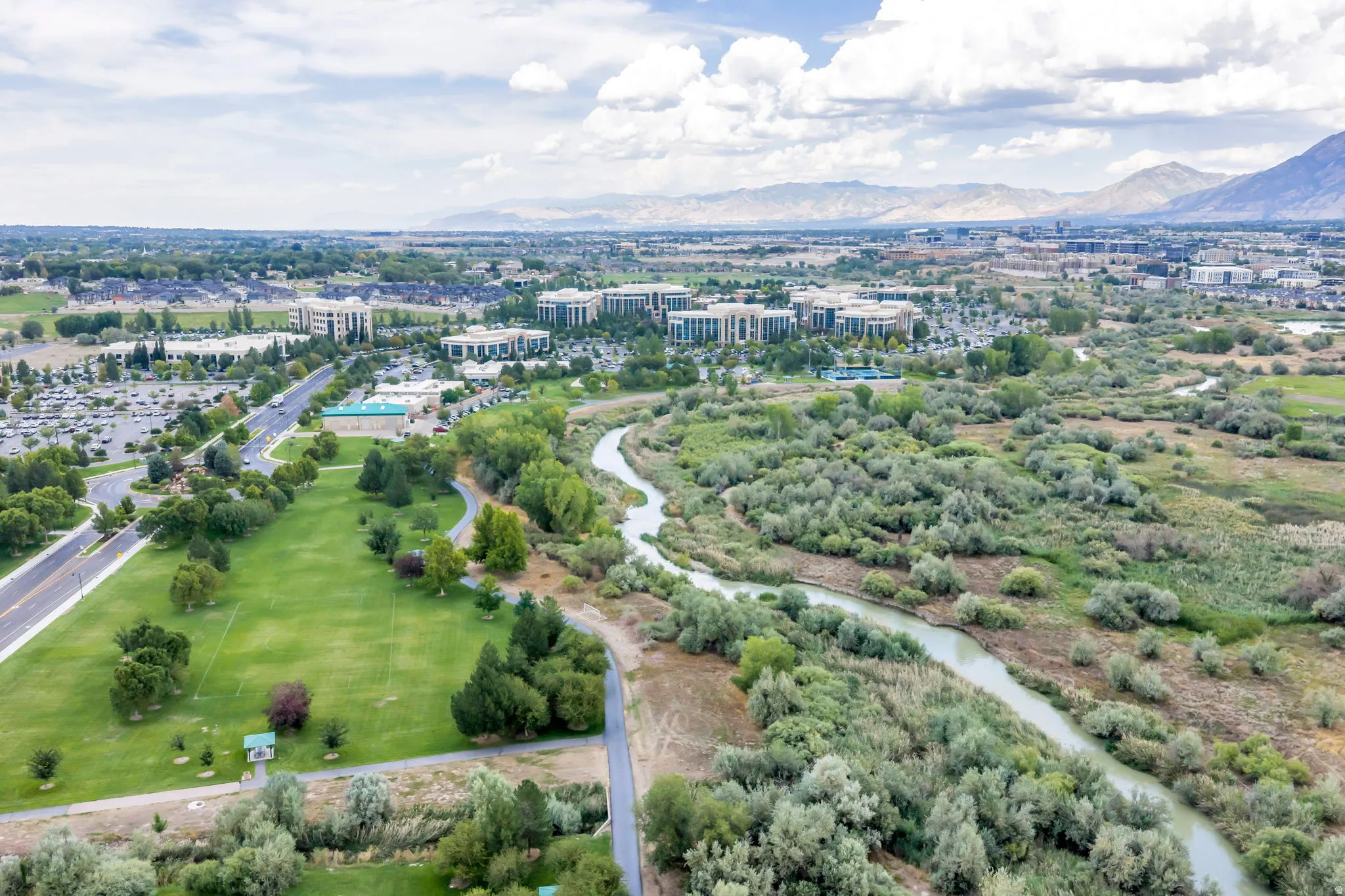 Aerial view showcasing expansive manicured grounds with mature tree-lined landscape, meandering waterway, and adjacent developed community with modern residential architecture set against mountain backdrop and verdant valley terrain
