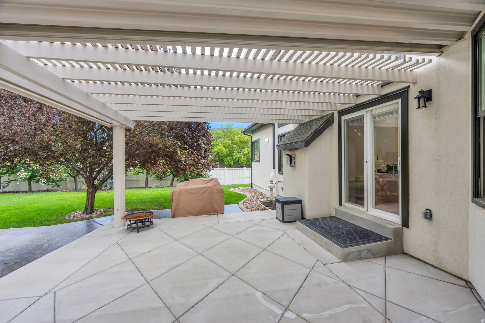 Expansive concrete patio with a white slatted pergola, overlooking a manicured lawn, with a sliding glass door entry to the interior