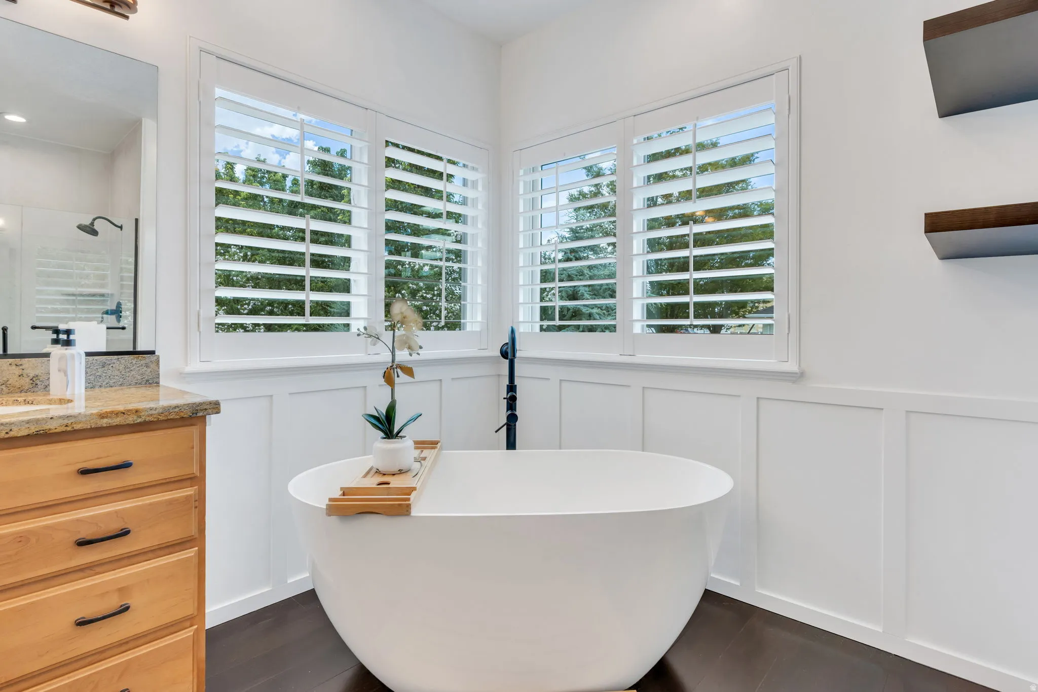 Freestanding soaking tub, white plantation shutters, rich wood-finish flooring, white wainscoting, and a light wood vanity with granite countertop