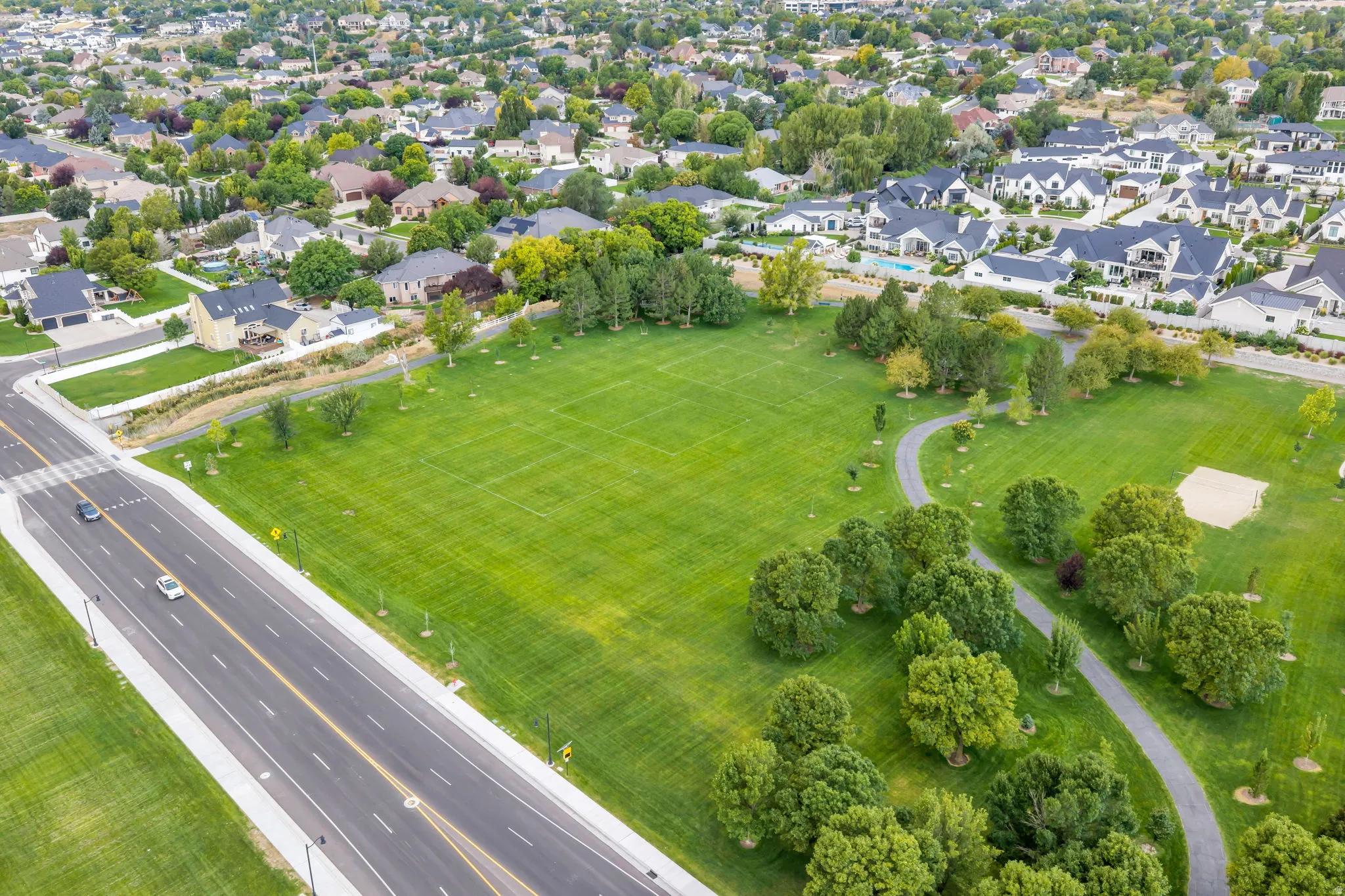 Expansive green space featuring a paved pathway, mature trees, and a sand court