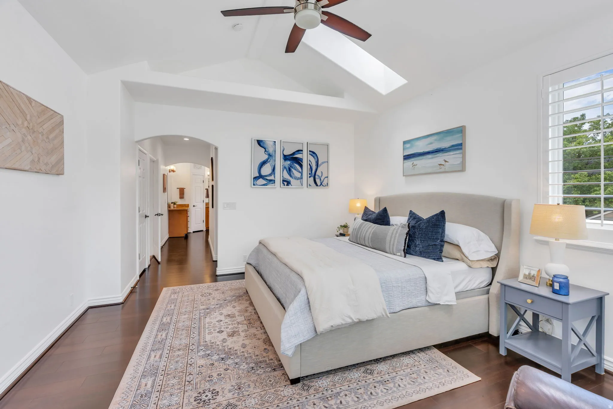 Vaulted ceiling with skylight, dark wood-finish flooring, ceiling fan, window with plantation shutters, and an arched doorway