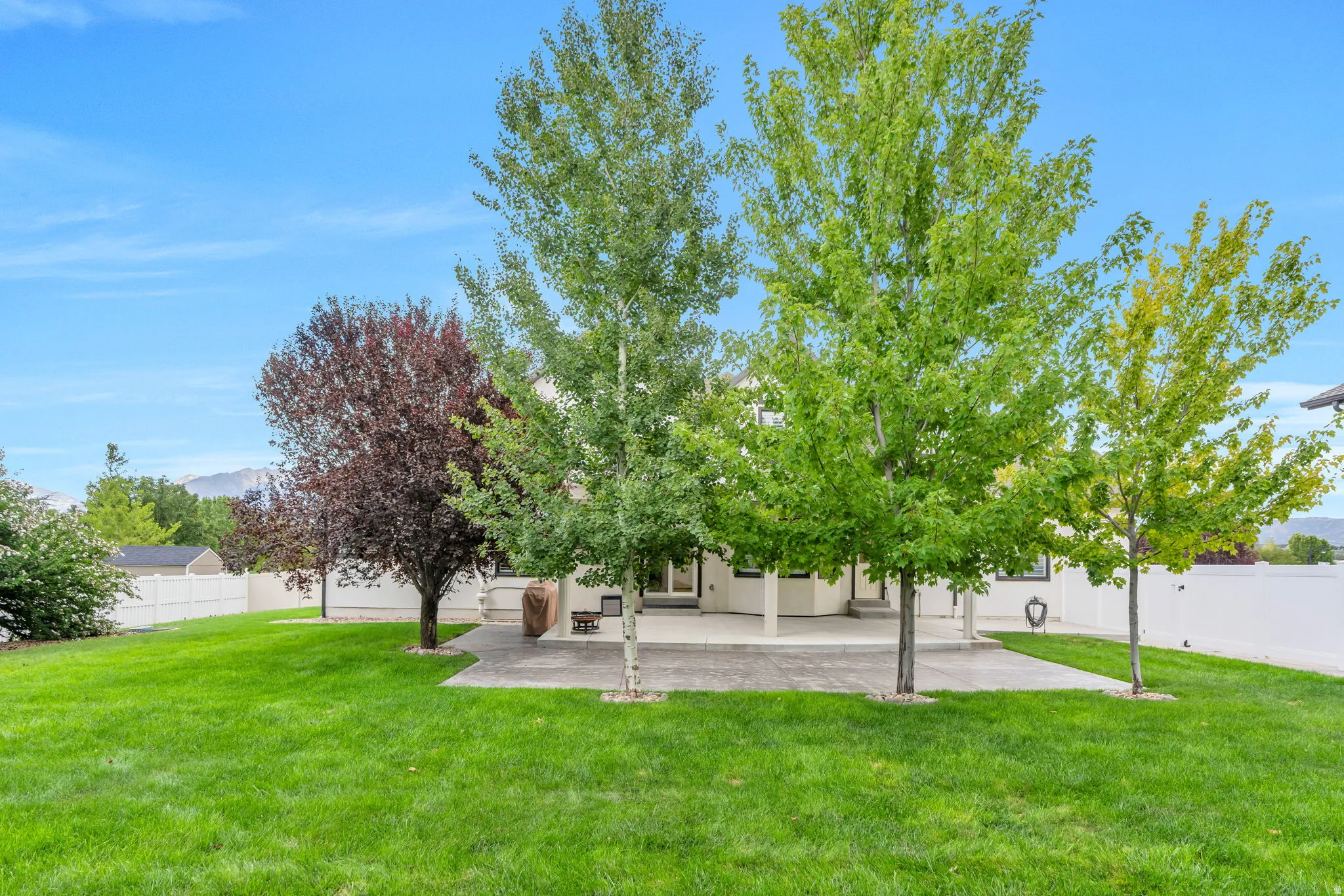Expansive green lawn with mature trees, a private white perimeter fence, and a covered concrete patio extending from the residence