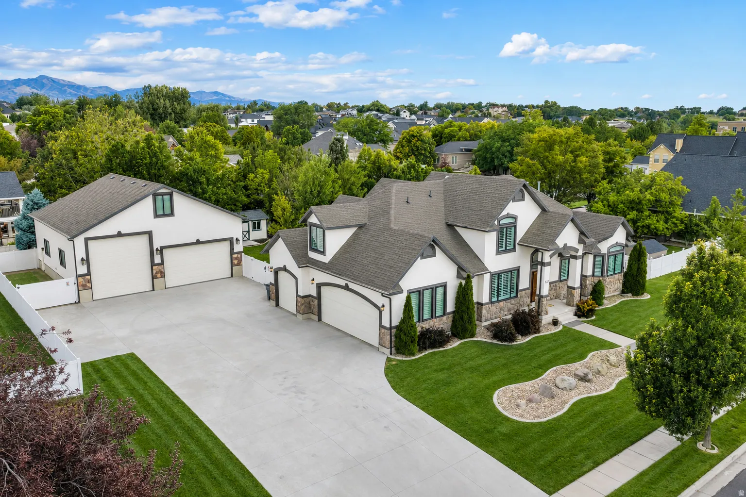 Impressive aerial view showcasing the home’s beautiful curb appeal, expansive driveway, attached garage, and oversized detached garage—surrounded by mature trees and a well-established neighborhood, with Wasatch Mountain views to the east.