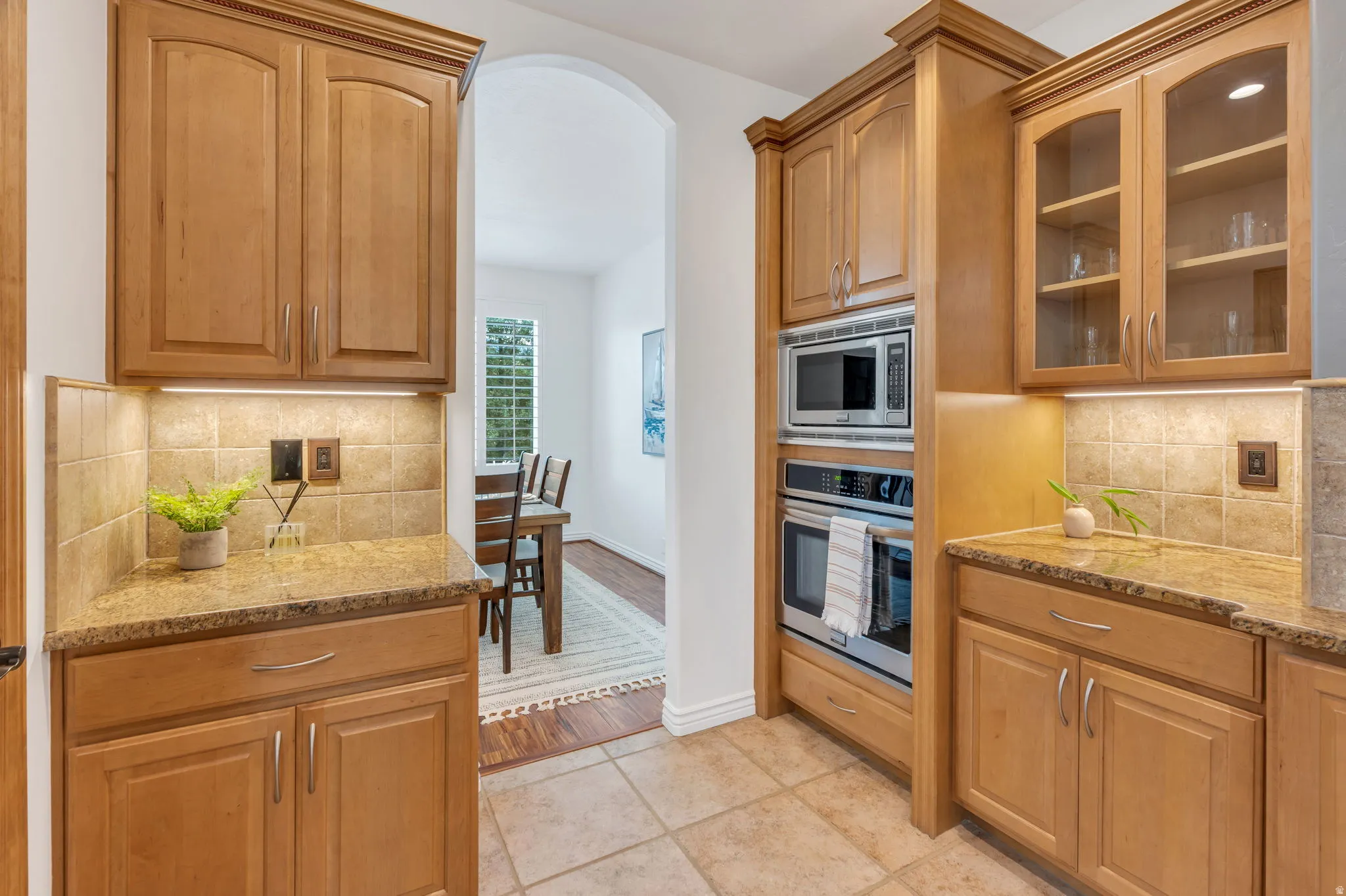 Light-toned wood cabinetry with glass-front uppers, stainless steel built-in microwave and oven, granite countertops, and tiled backsplash