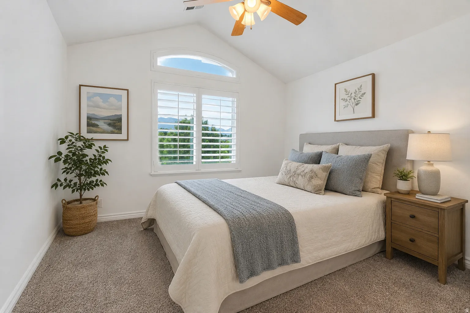 Bright primary bedroom showcasing vaulted ceilings with ceiling fan, expansive transom window with plantation shutters, neutral bedding, wood-finish nightstands with integrated lighting, and soft carpet flooring throughout