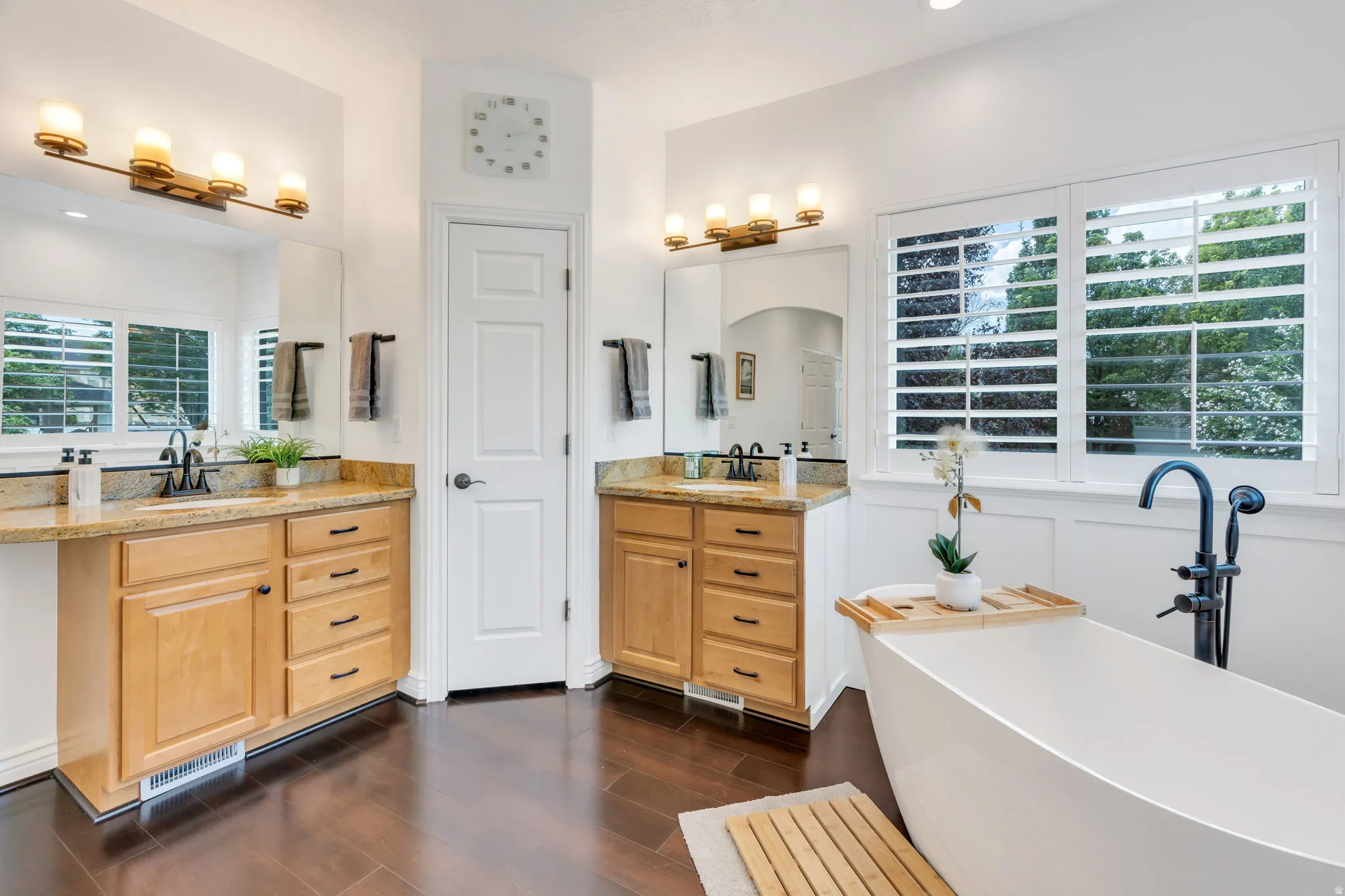 Freestanding soaking tub with floor-mounted fixture, dual vanity with undermount sinks, wood-finish flooring, and shuttered windows