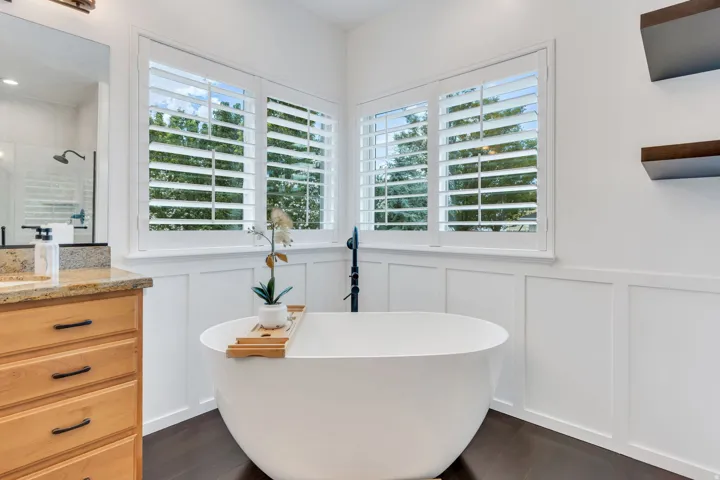 Freestanding soaking tub, white plantation shutters, rich wood-finish flooring, white wainscoting, and a light wood vanity with granite countertop
