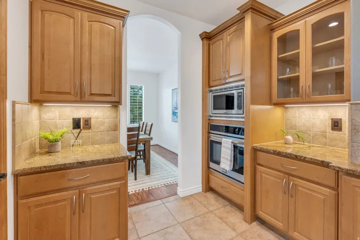 Light-toned wood cabinetry with glass-front uppers, stainless steel built-in microwave and oven, granite countertops, and tiled backsplash