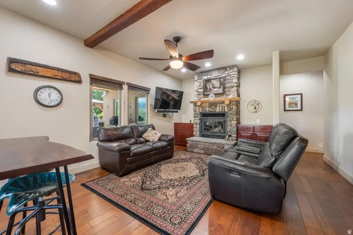 Living area featuring a stone-clad fireplace, wood-finish flooring, exposed ceiling beams, recessed lighting, and a ceiling fan