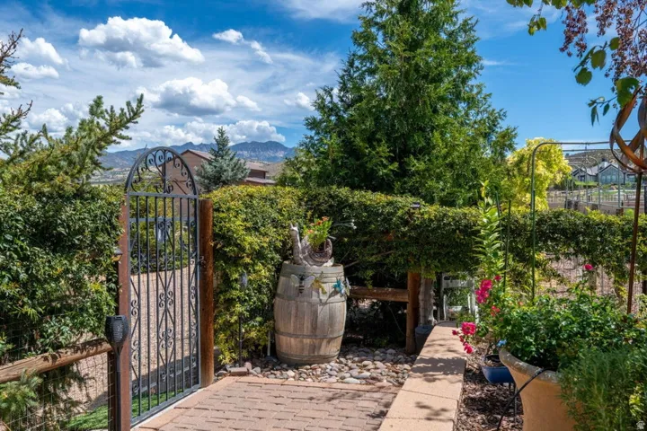 Wrought iron gate with decorative archway, brick paver walkway, lush privacy hedging, and a rustic wooden barrel accent