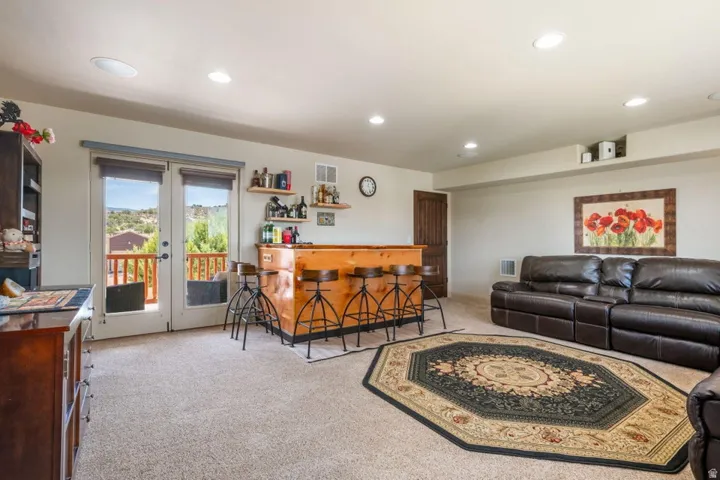 Built-in wooden bar with integrated shelving, light-colored carpeting, recessed ceiling lighting, and French doors leading to an outdoor balcony with a wooden railing