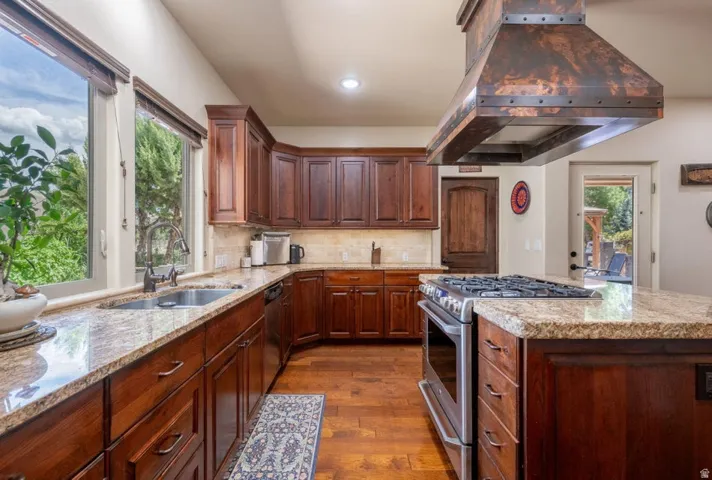 Kitchen featuring wood-finish cabinetry, light-toned stone countertops, stainless steel appliances, a center island with a gas range, and a decorative range hood