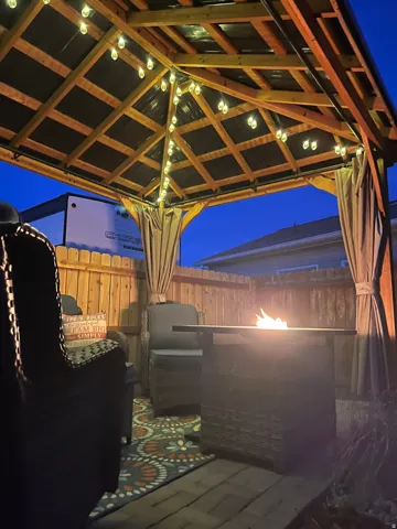 Wood-framed gazebo featuring string lighting, privacy drapes, and a stone-finish fire pit