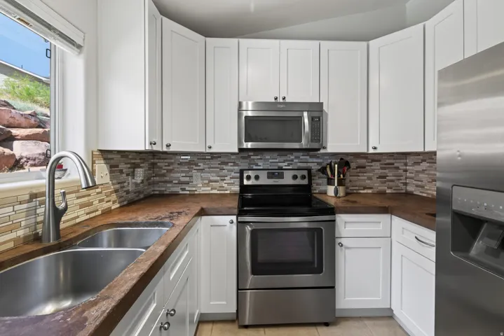 White shaker-style cabinetry, stainless steel appliances, mosaic tile backsplash, double basin stainless steel sink, and wood-finish countertops