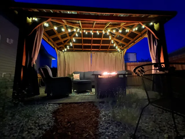 Outdoor gazebo with a slatted wood roof, string lighting, and privacy curtains