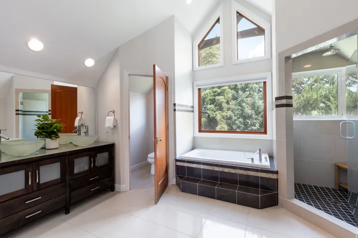 Dual vessel sinks, expansive vanity with dark wood-finish cabinetry, soaking tub with tile surround, walk-in shower with glass enclosure, and multiple windows