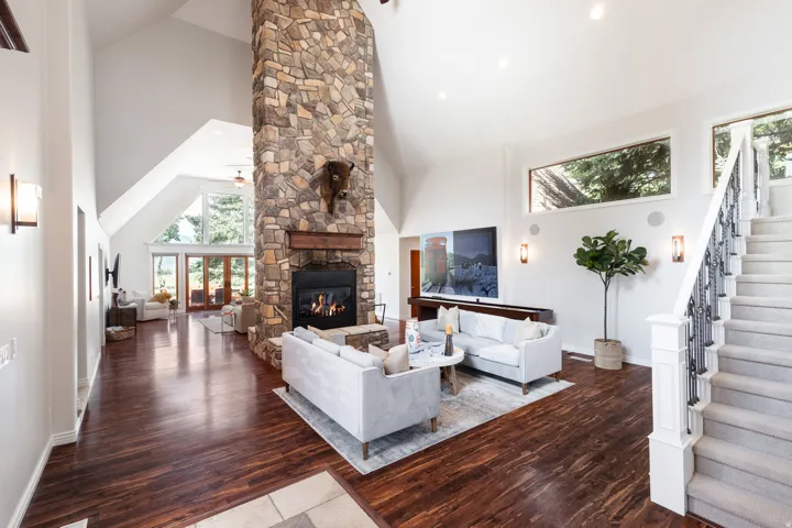 Vaulted ceilings with abundant natural light, a stone-clad fireplace with wood mantel, wood-finish flooring, and a carpeted staircase with an ornamental wrought iron railing