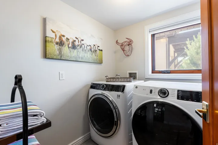 Dedicated laundry room featuring side-by-side front-load washers and dryers with digital controls, ample counter space for folding, natural light from a large window, and neutral wall finishes for a clean, functional workspace