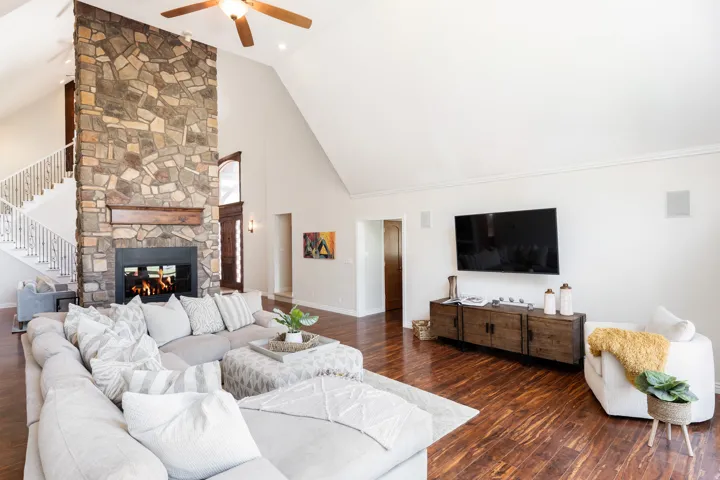 Vaulted ceilings, a floor-to-ceiling stone fireplace with a wood mantle, wood-finish flooring, a ceiling fan, and built-in wall speakers