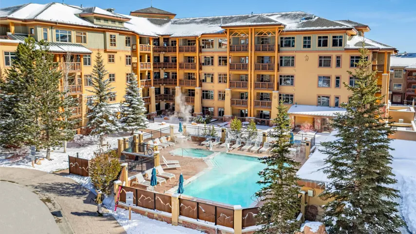 Multi-story building with a snow-covered roof, multiple balconies, a swimming pool, and an outdoor hot tub surrounded by evergreen trees