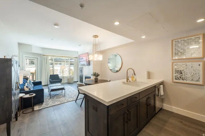 Open-concept living area with wood-finish flooring, a kitchen island featuring a deep undermount sink and a brass-finish faucet, recessed lighting, and a large window