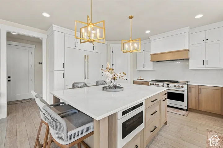 Spacious kitchen featuring an expansive central island with a built-in microwave, white cabinetry, wood-finish base cabinets, and matching wood-finish flooring. Stunning Matt white and gold fridge, dishwasher and range.