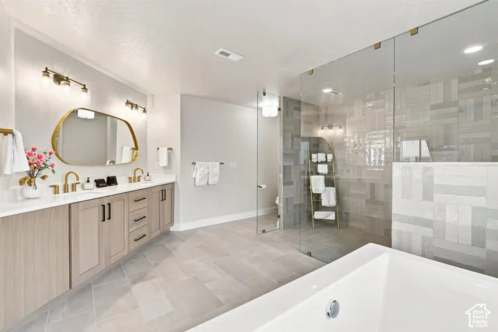 Primary Bathroom. beautiful tile work. Dual vanity with light wood-finish cabinetry, white countertop, and gold-finish fixtures and ceiling to floor glass enclosure for shower.