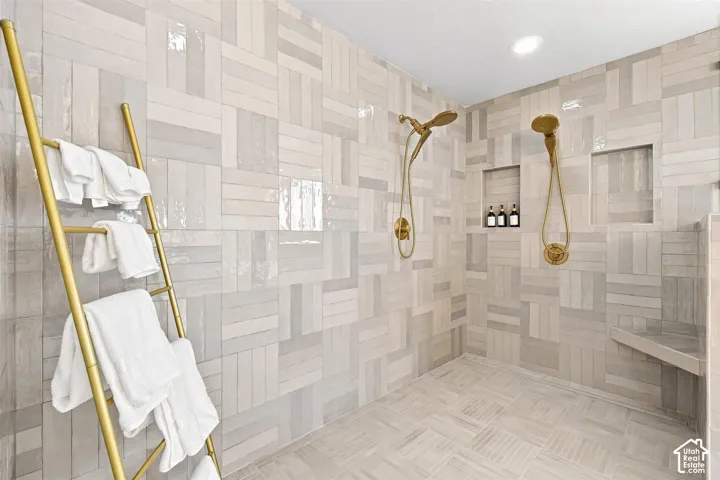 Spacious walk-in shower featuring patterned wall and floor tiling, dual shower heads with gold-tone fixtures, two recessed shelving niches, and a built-in shower bench. Floor to ceiling glass enclosure.