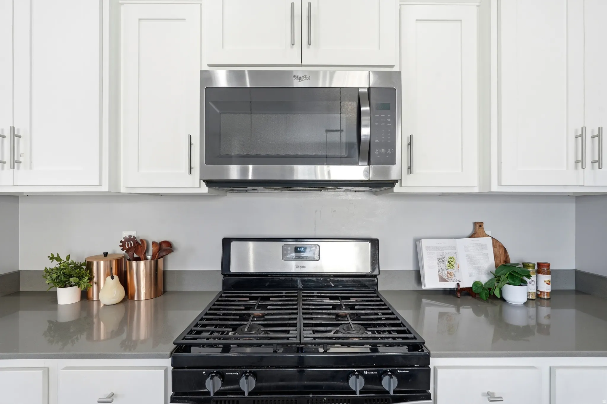 Stainless steel microwave, white shaker-style cabinetry with silver hardware, smooth grey countertops, and a black gas range